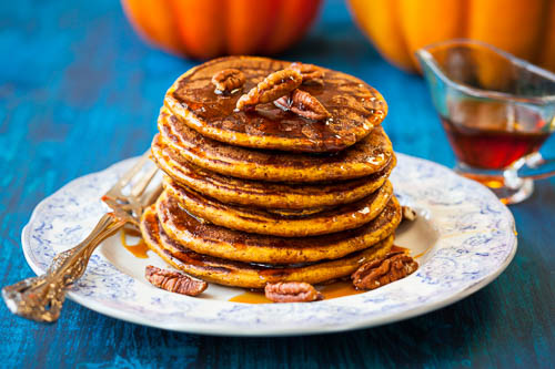 A stack of butternut squash pancakes on a white plate with a blue tablecloth, topped with syrup and pecan nuts