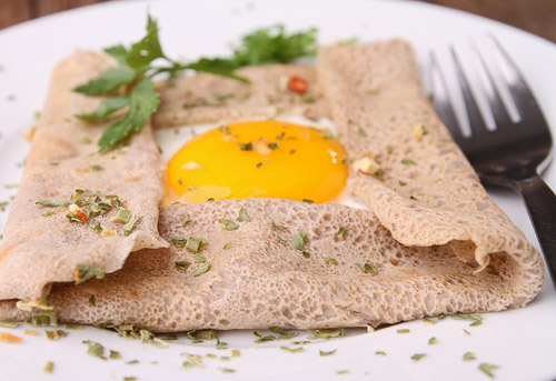 Closeup of a dairy-free buckwheat pancake loaded with a savory egg and topped with spices and green herbs.