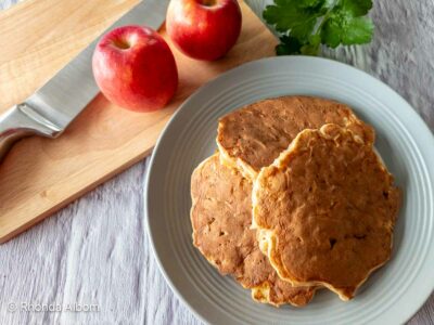 Gluten free apple pancakes on a plate next to apples on a chopping board with the knife