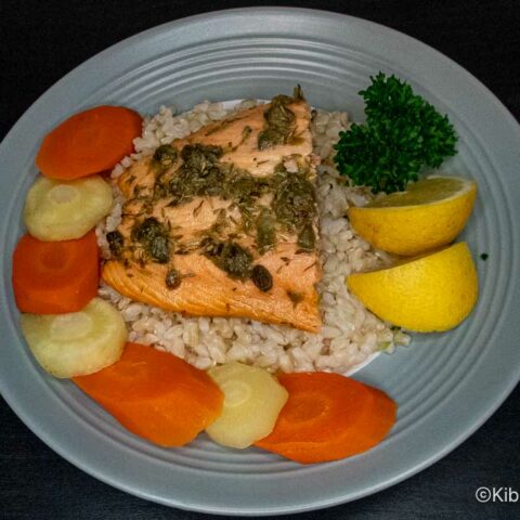 Plate of baked salmon with capers on a bed of brown rice with carrots, parsnips and lemon wedges.